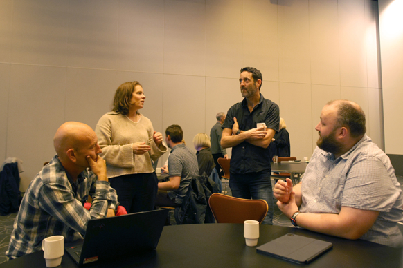 To personer står og to sitter ved bordet ved sinden av