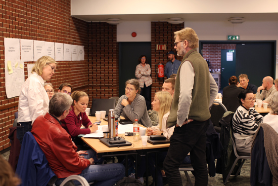 Flere personer sitter rundt et bord og diskuterer. Noen står.