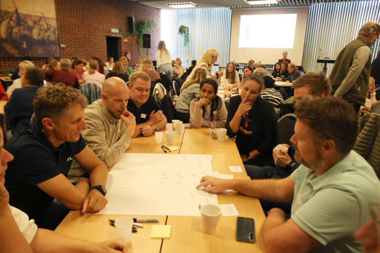 Flere personer sitter rundt et stort bord. De peker og ser på en plakat på bordet hvor det står ulike gevinster.