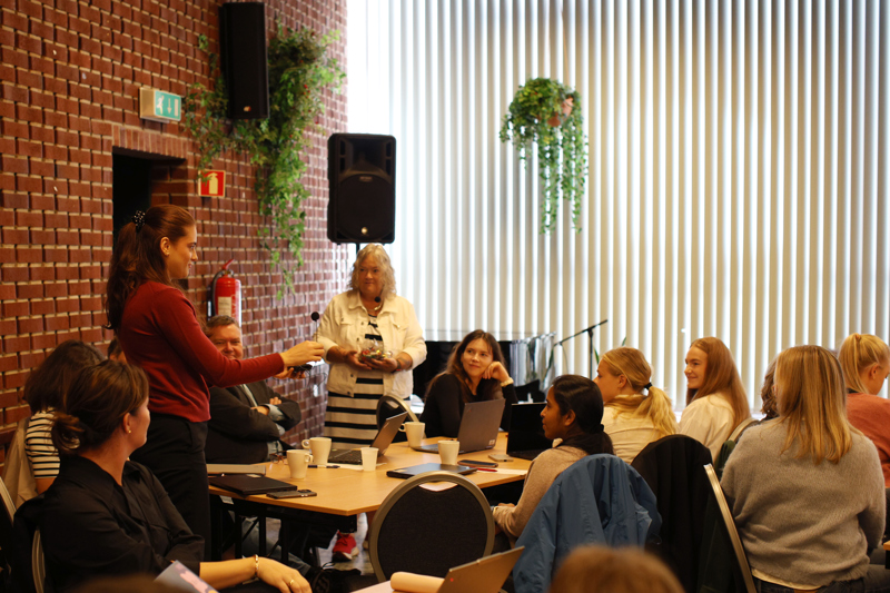 En rekke folk sitter rundt et bord i den store salen. En står og snakker. 