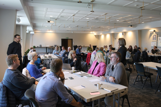 Salen med alle deltakerne diskuterer rundt bordene. Arve Ubøe Rohde og Ruth Økland står ved et av de fremste bordene.