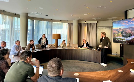 Deltakerne på workshopen sitter rundt det runde bordet. Haglund snakker og står foran skjermen med presentasjonen på