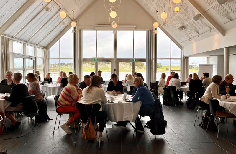 Bildet viser hele lokalet hvor møtet ble holdt. Vi ser mange runde bord, hvor det sitter folk rundt som snakker med hverandre.