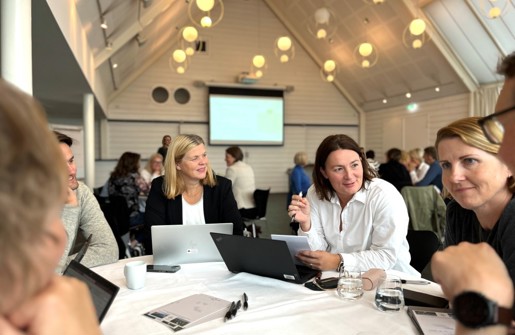 Bildet viser et av bordene i møterommet på samlingen, hvor Kristine Skjøthaug og Trine Voos sitter i livlig prat med de andre rundt bordet.