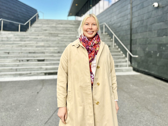 Vi ser prosjektleder Ellen Bowitz som står utenfor et stort bygg i solen. Bygget er i grå skifer, Herbarium. Hun smiler og er kledd i lys kåpe.