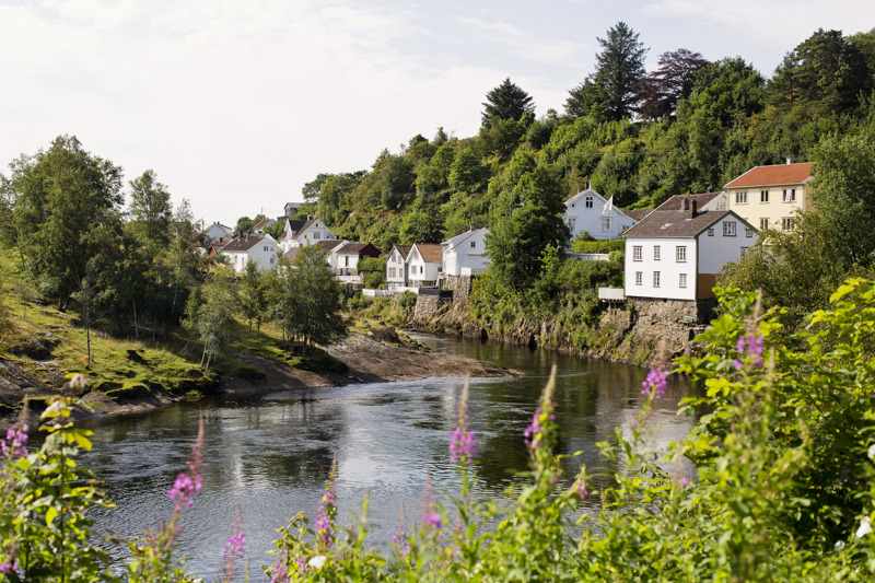 Vi ser koselige små trehus i Sogndalstrand, som ligger ved en elv. Det er sommer, grønne busker og trær.