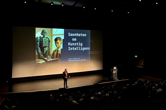Bildet viser Morten Goodwin på scenen og folk som sitter i salen. På den store skjermen bak han står det "sannheten om kunstig intelligens".